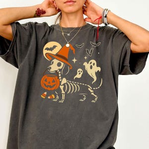 May include: Dark gray t-shirt featuring a Halloween-themed graphic. The design includes a skeleton dachshund wearing a witch's hat and carrying a jack-o'-lantern. Bats, ghosts, and a full moon complete the spooky illustration.