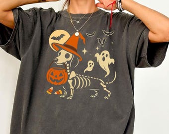 May include: Dark gray t-shirt featuring a Halloween-themed graphic. The design includes a skeleton dachshund wearing a witch's hat and carrying a jack-o'-lantern. Bats, ghosts, and a full moon complete the spooky illustration.
