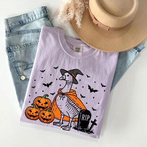 Puede incluir: Una camiseta morada con un dibujo animado de un ganso con un sombrero de bruja y una capa. El ganso est&aacute; de pie junto a una l&aacute;pida que dice "RIP" y tres calabazas. Hay murci&eacute;lagos y estrellas en el fondo.