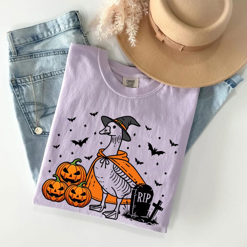 Puede incluir: Una camiseta morada con un dibujo animado de un ganso con un sombrero de bruja y una capa. El ganso est&aacute; de pie junto a una l&aacute;pida que dice "RIP" y tres calabazas. Hay murci&eacute;lagos y estrellas en el fondo.