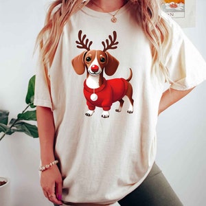 May include: A white t-shirt with a cartoon image of a dachshund wearing a red sweater and reindeer antlers. The dachshund has a red nose and brown and white fur.