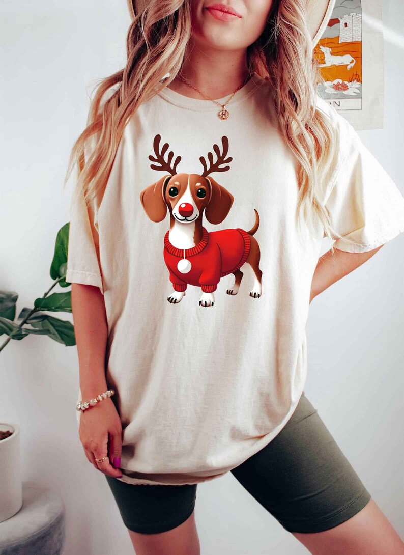 May include: A white t-shirt with a cartoon image of a dachshund wearing a red sweater and reindeer antlers. The dachshund has a red nose and brown and white fur.