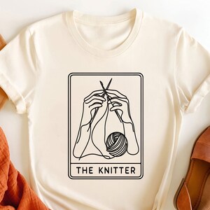May include: A white t-shirt featuring a black line drawing of hands knitting a ball of yarn. The design is within a rectangle with the text "THE KNITTER" underneath.