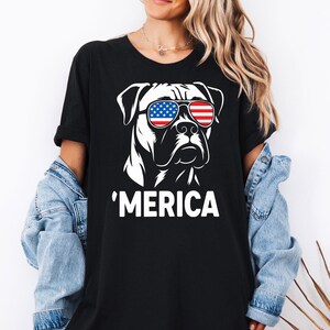 May include: Black t-shirt with a white graphic of a dog wearing sunglasses in the style of the American flag. The word "'MERICA" is printed in white below the dog illustration. The t-shirt is a casual style.