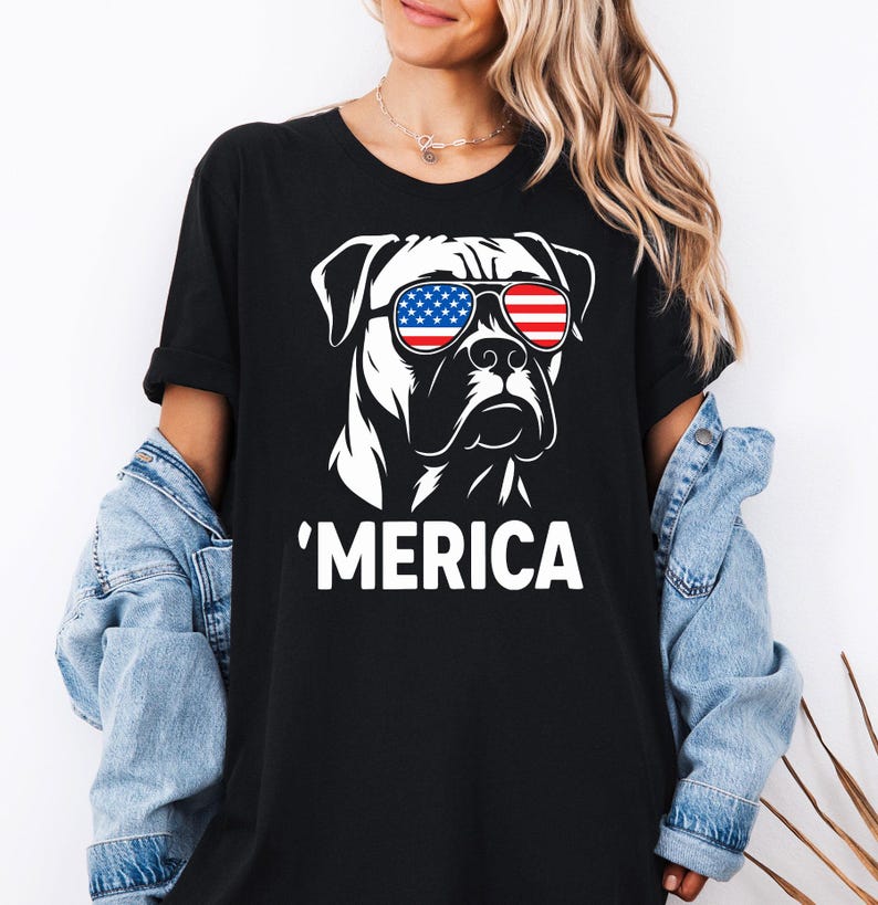 May include: Black t-shirt with a white graphic of a dog wearing sunglasses in the style of the American flag. The word "'MERICA" is printed in white below the dog illustration. The t-shirt is a casual style.