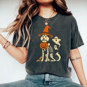 May include: Dark gray t-shirt featuring a Halloween-themed graphic. The design includes a dog wearing a witch's hat and holding a pumpkin, with a skeleton body and ghosts. The shirt is a comfortable, casual style.