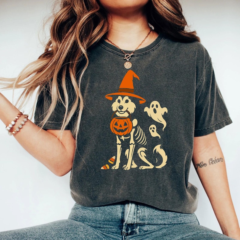 May include: Dark gray t-shirt featuring a Halloween-themed graphic. The design includes a dog wearing a witch's hat and holding a pumpkin, with a skeleton body and ghosts. The shirt is a comfortable, casual style.
