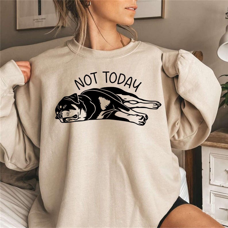 May include: Beige sweatshirt with the text "NOT TODAY" above a black and white graphic of a dog lying down. The dog graphic is a Rottweiler breed. The sweatshirt has long sleeves and a crew neck.