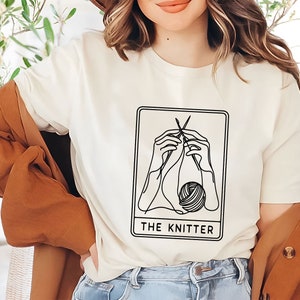 May include: White t-shirt featuring a black line drawing of hands knitting a ball of yarn. The design is within a card-like frame with the text "THE KNITTER" underneath.