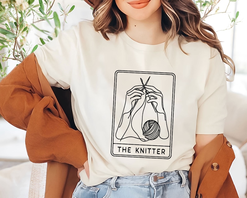 May include: White t-shirt featuring a black line drawing of hands knitting a ball of yarn. The design is within a card-like frame with the text "THE KNITTER" underneath.