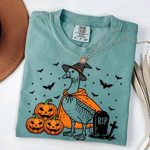 Puede incluir: Una camiseta azul turquesa con un dibujo animado de un ganso con un sombrero de bruja y una capa. El ganso est&aacute; de pie junto a tres calabazas y una l&aacute;pida con el texto "RIP".