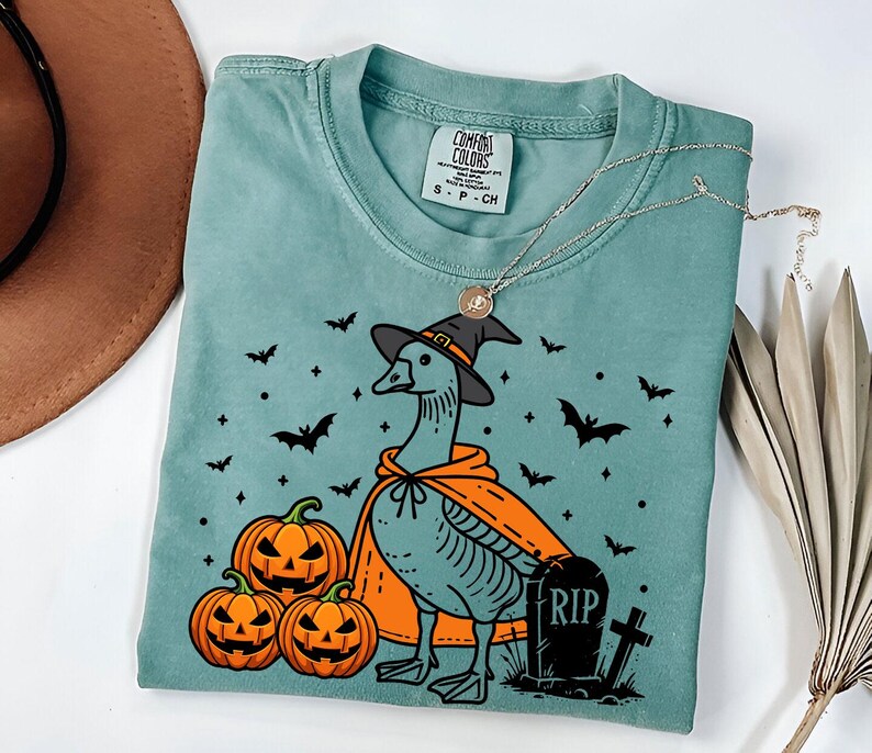 Puede incluir: Una camiseta azul turquesa con un dibujo animado de un ganso con un sombrero de bruja y una capa. El ganso est&aacute; de pie junto a tres calabazas y una l&aacute;pida con el texto "RIP".