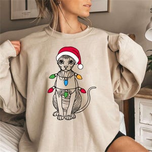 May include: A beige sweatshirt with a graphic of a cat wearing a Santa hat and Christmas lights. The cat is a Sphynx breed with no fur.