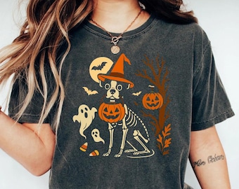 Puede incluir: Camiseta gris oscuro con un diseño de Halloween. El diseño incluye un esqueleto de perro con un sombrero de bruja y una cabeza de calabaza, con fantasmas, murciélagos y una luna llena. La camiseta tiene cuello redondo y mangas cortas.