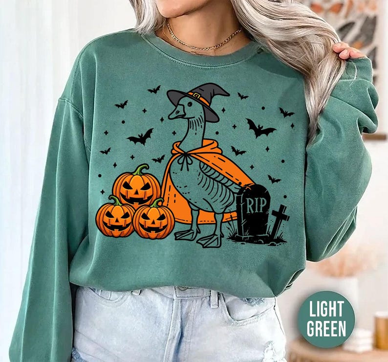 May include: A light green sweatshirt with a graphic of a goose wearing a witch hat and a cape. The goose is surrounded by pumpkins and a tombstone that says "RIP".