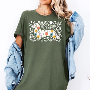 May include: Olive green t-shirt featuring a floral dachshund design. The dog is filled with colorful flowers, surrounded by white floral accents. The shirt has short sleeves and a relaxed fit.