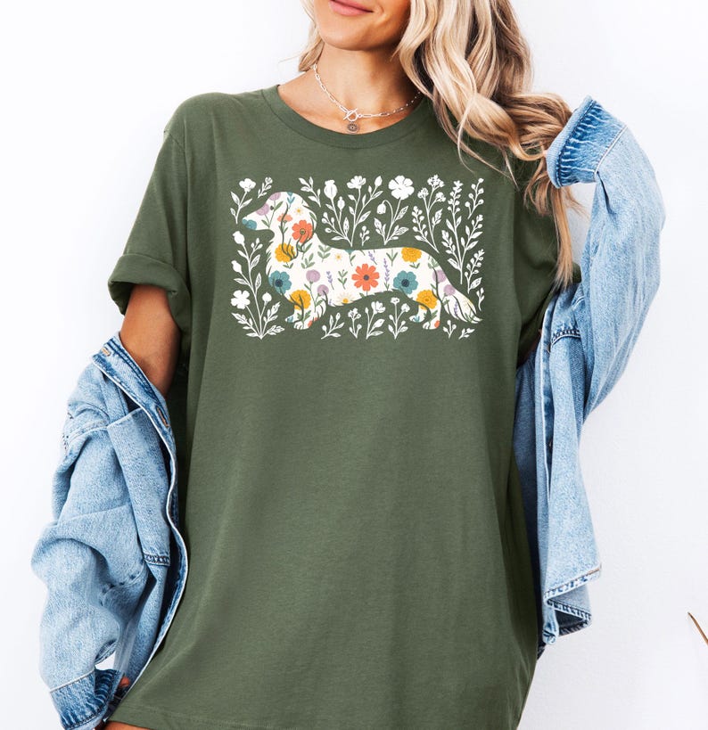 May include: Olive green t-shirt featuring a floral dachshund design. The dog is filled with colorful flowers, surrounded by white floral accents. The shirt has short sleeves and a relaxed fit.