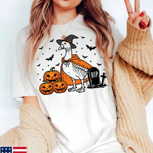 Puede incluir: Camiseta blanca con un gr&aacute;fico de un ganso con un sombrero de bruja y una capa, sosteniendo una calabaza. Hay murci&eacute;lagos y una l&aacute;pida con el texto "RIP" en ella.