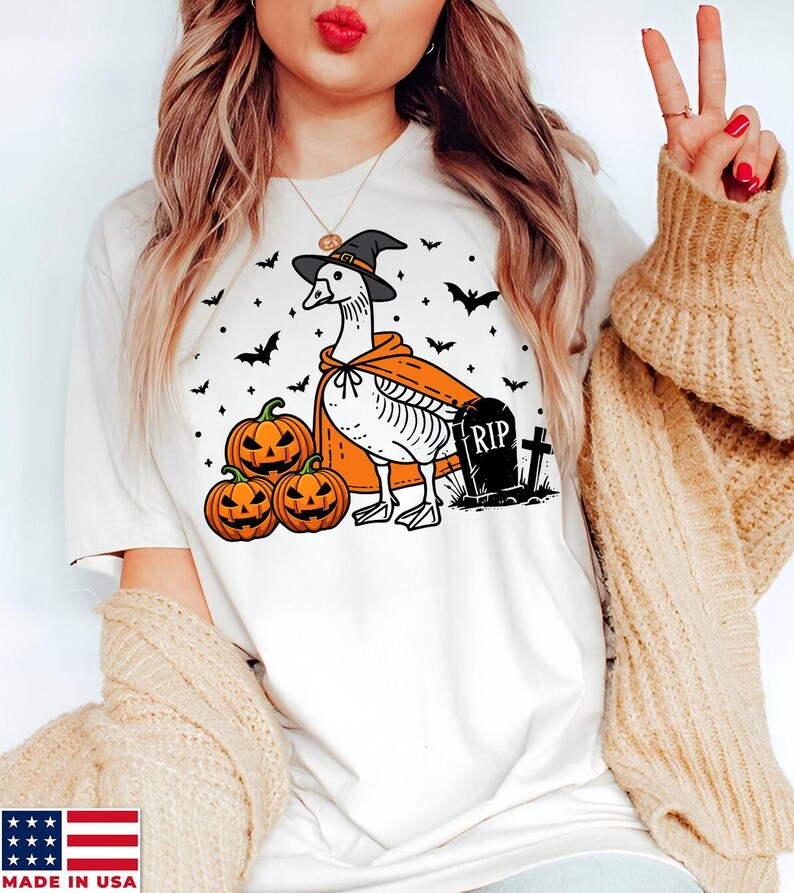 Puede incluir: Camiseta blanca con un gr&aacute;fico de un ganso con un sombrero de bruja y una capa, sosteniendo una calabaza. Hay murci&eacute;lagos y una l&aacute;pida con el texto "RIP" en ella.