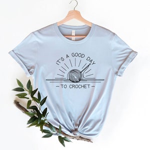 May include: A light blue t-shirt with a black graphic of a yarn ball and crochet hook in a sun design. The text reads "It's a good day to crochet."