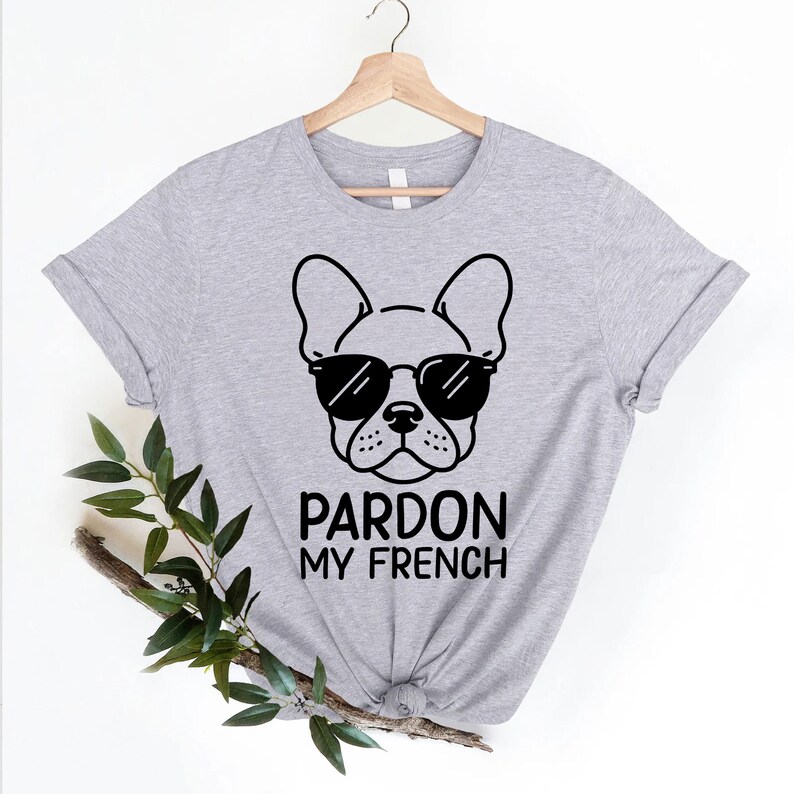 K&ouml;nnte beinhalten: Graues T-Shirt mit einer schwarz-wei&szlig;en Illustration eines franz&ouml;sischen Bulldoggen mit Sonnenbrille. Der Text "Pardon My French" ist unter dem Bild gedruckt.