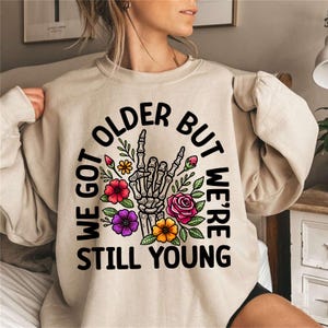 May include: A beige sweatshirt with a black graphic print that says "We got older but we're still young". The graphic features a skeleton hand making a rock and roll sign with flowers around it.