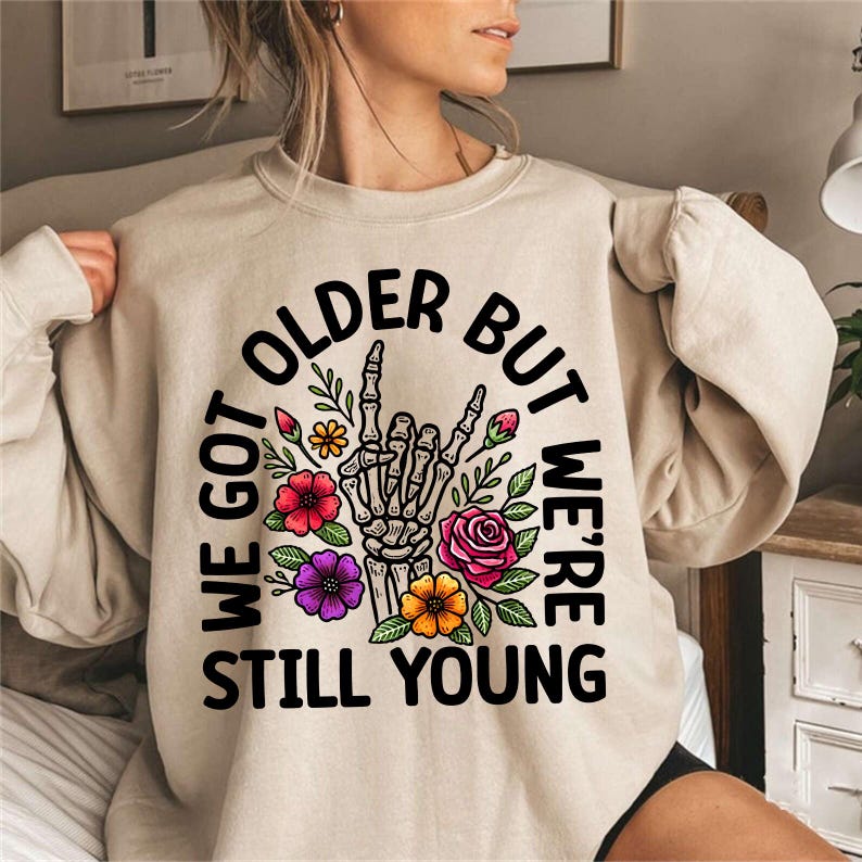 May include: A beige sweatshirt with a black graphic print that says "We got older but we're still young". The graphic features a skeleton hand making a rock and roll sign with flowers around it.