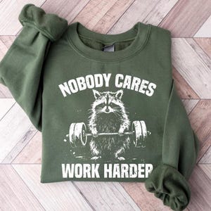 May include: Green sweatshirt with a white graphic of a raccoon lifting weights. The text "Nobody Cares Work Harder" is printed on the sweatshirt.