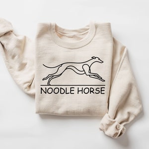 May include: A beige sweatshirt with a black line drawing of a greyhound dog running and the text "NOODLE HORSE" below the dog.