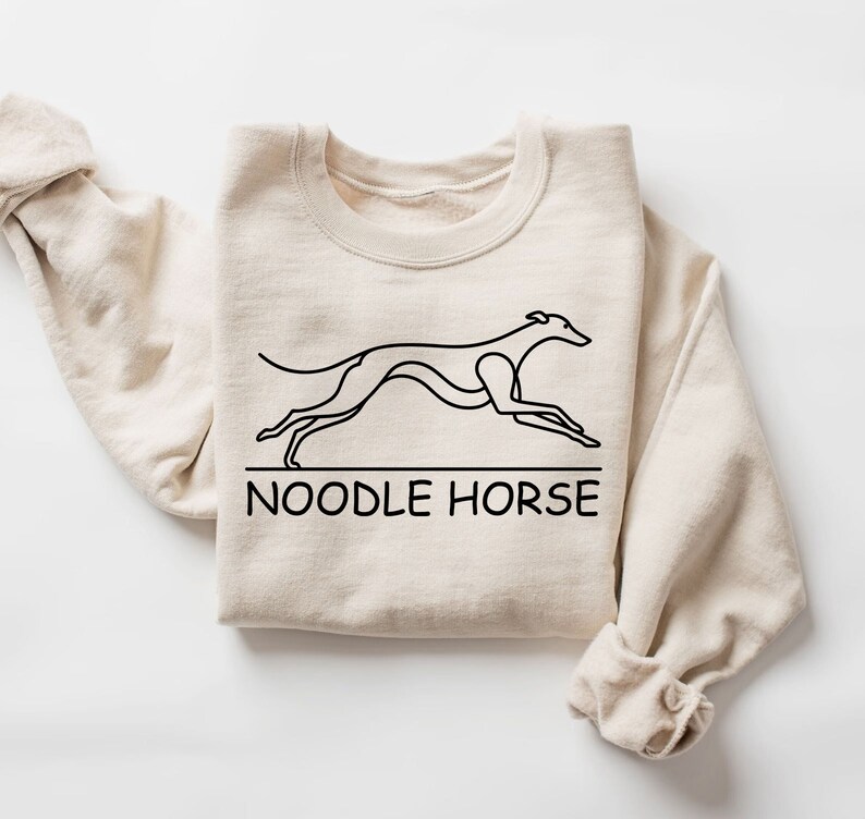 May include: A beige sweatshirt with a black line drawing of a greyhound dog running and the text "NOODLE HORSE" below the dog.