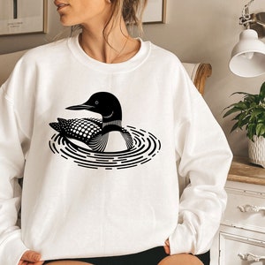 May include: A white sweatshirt with a black and white graphic of a loon swimming in a circle of ripples.