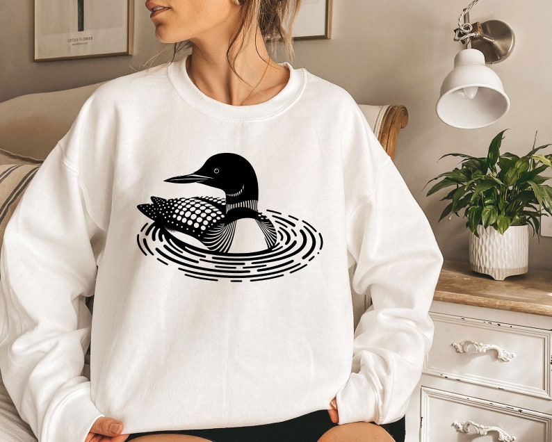 May include: A white sweatshirt with a black and white graphic of a loon swimming in a circle of ripples.