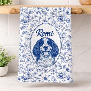 Pode incluir: Toalha de cozinha branca com um padrão floral azul e um oval central com um retrato de cão e o nome "Remi". O cão é retratado em azul e branco, com uma expressão feliz.