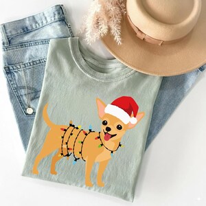 May include: A light green t-shirt featuring a cartoon chihuahua wearing a Santa hat and string lights. The dog is tan with a red and white hat and a black string of colorful lights. The shirt is folded on top of blue jeans and a tan hat.