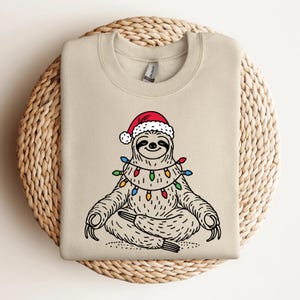 Könnte beinhalten: Ein beiger Pullover mit einer Cartoon-Illustration eines Faultiers, das einen Weihnachtsmann-Hut und Weihnachtslichter trägt. Das Faultier sitzt in einer Yoga-Pose.