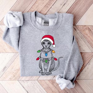 May include: A gray sweatshirt with a black and white illustration of a cat wearing a red Santa hat and Christmas lights.