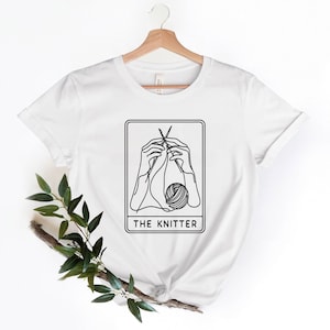 May include: White t-shirt featuring a black line drawing of hands knitting with yarn and needles. The drawing is enclosed in a black rectangle with the text "THE KNITTER" beneath it.