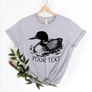 May include: A gray t-shirt with a black and white illustration of a loon swimming in water. The text "YOUR TEXT" is printed below the loon.