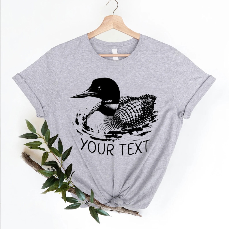 May include: A gray t-shirt with a black and white illustration of a loon swimming in water. The text "YOUR TEXT" is printed below the loon.