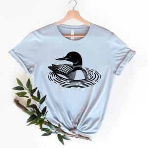 May include: A light blue t-shirt with a black and white graphic of a loon swimming in a circle of ripples.