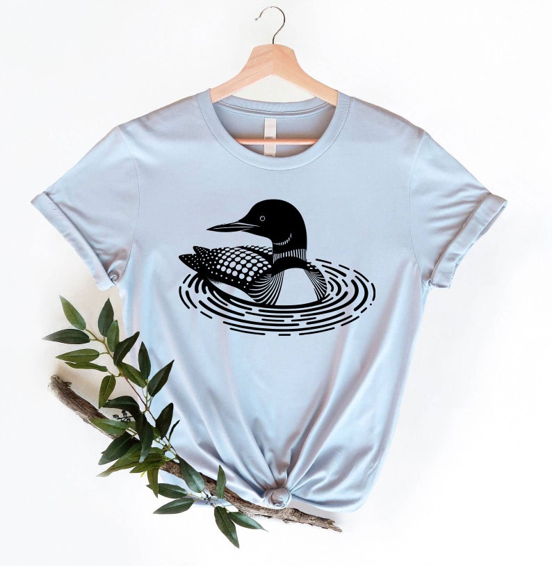 May include: A light blue t-shirt with a black and white graphic of a loon swimming in a circle of ripples.