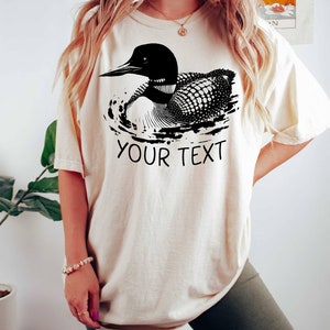 May include: A cream colored t-shirt with a black and white illustration of a loon swimming. The text "YOUR TEXT" is printed below the loon.