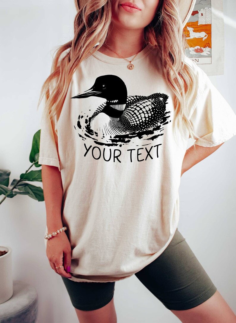 May include: A cream colored t-shirt with a black and white illustration of a loon swimming. The text "YOUR TEXT" is printed below the loon.