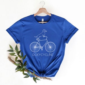 May include: A royal blue t-shirt with a white outline of a dachshund riding a bicycle. The text "DOXYCYCLINE" is printed below the bicycle.