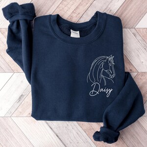 May include: A navy blue sweatshirt with a white outline of a horse's head and the name "Daisy" written in cursive below it.