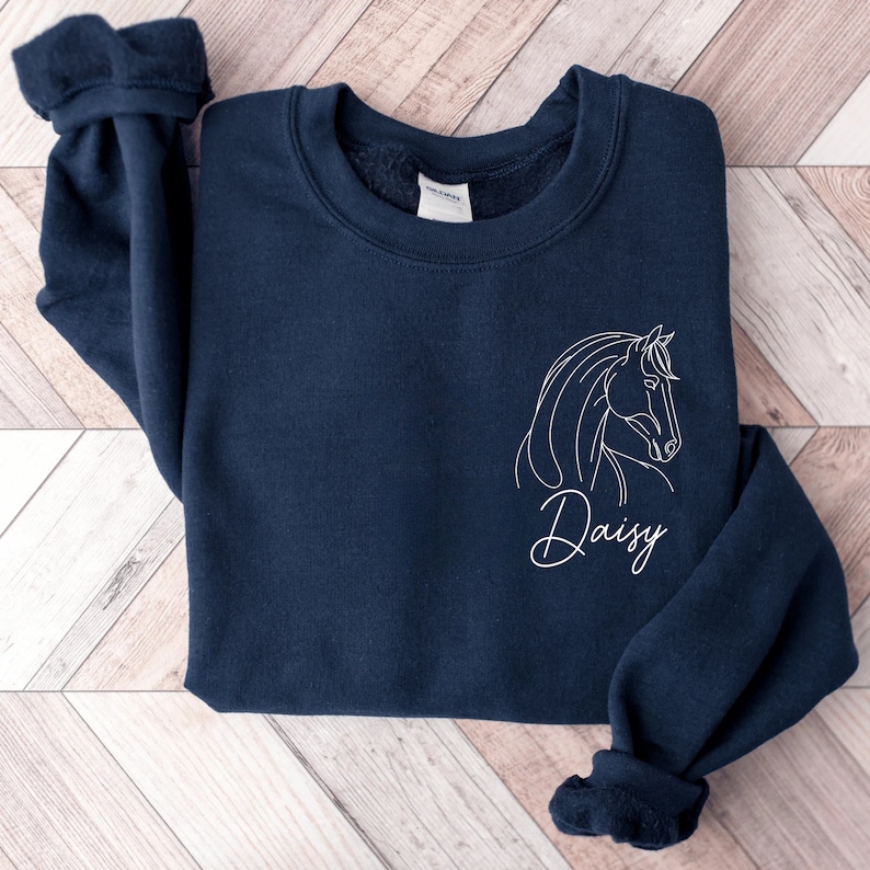 May include: A navy blue sweatshirt with a white outline of a horse's head and the name "Daisy" written in cursive below it.