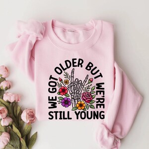 May include: Pink sweatshirt with a graphic print of a skeleton hand with flowers and the text "We got older but we're still young".
