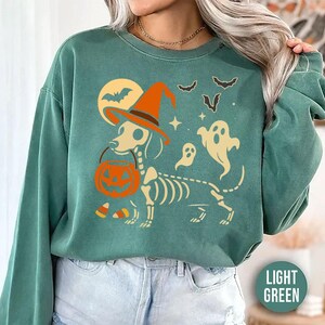 May include: Light green sweatshirt with a Halloween-themed graphic. The design features a dachshund skeleton wearing a witch hat and holding a pumpkin bucket. Bats, ghosts, and a full moon complete the spooky scene.