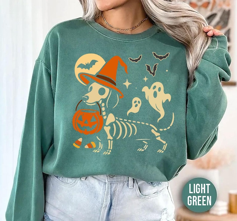 May include: Light green sweatshirt with a Halloween-themed graphic. The design features a dachshund skeleton wearing a witch hat and holding a pumpkin bucket. Bats, ghosts, and a full moon complete the spooky scene.