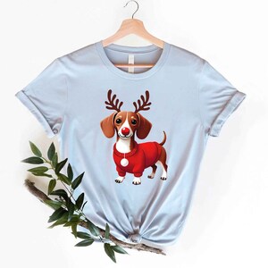 May include: A light blue t-shirt with a cartoon image of a dachshund wearing a red sweater and reindeer antlers. The dachshund has a red nose and white markings on its face.
